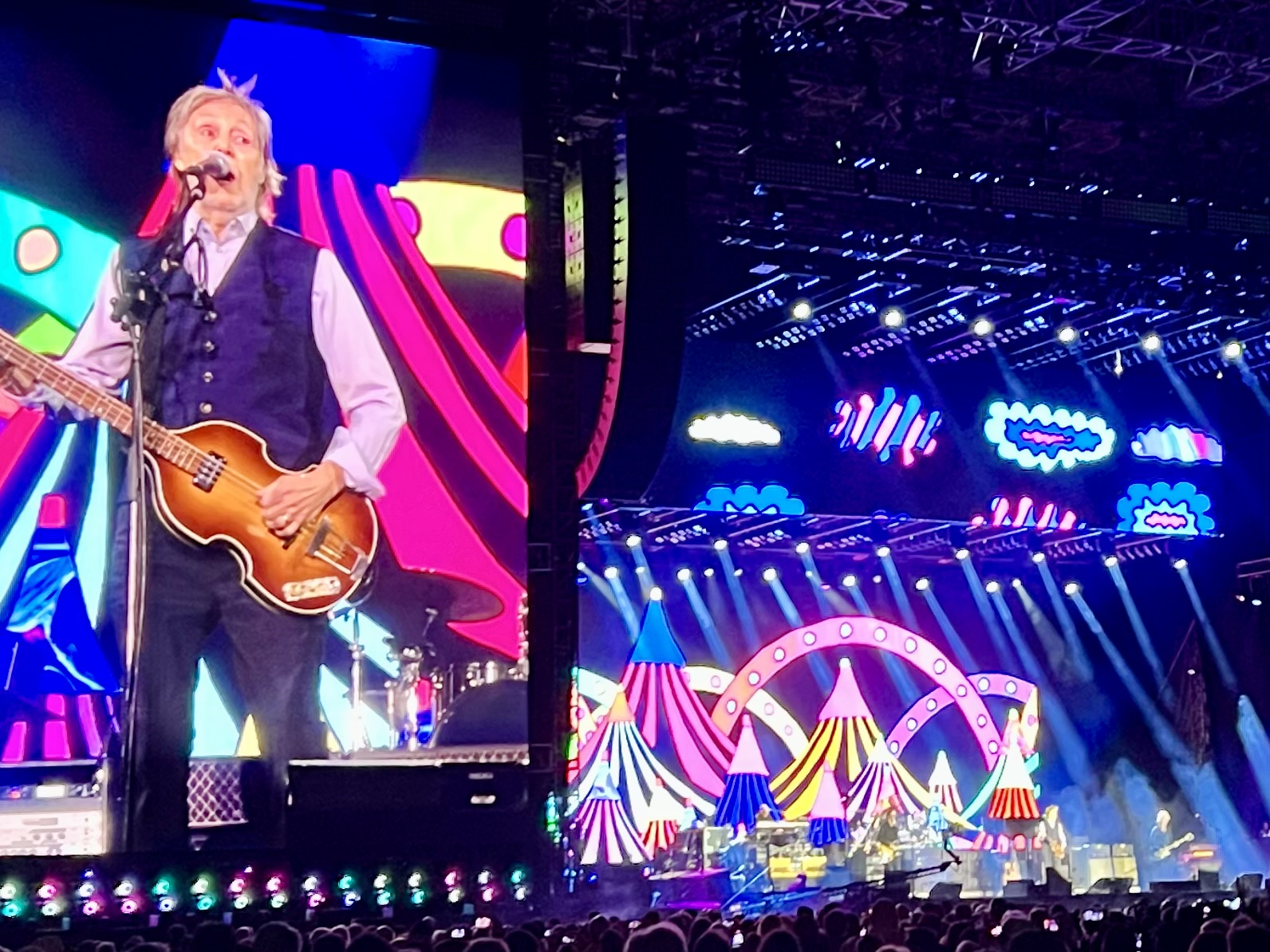 Paul McCartney performing on stage at Coors Field in Denver on a color and bright stage, including a large projection of him playing his trademark Hofner bass.