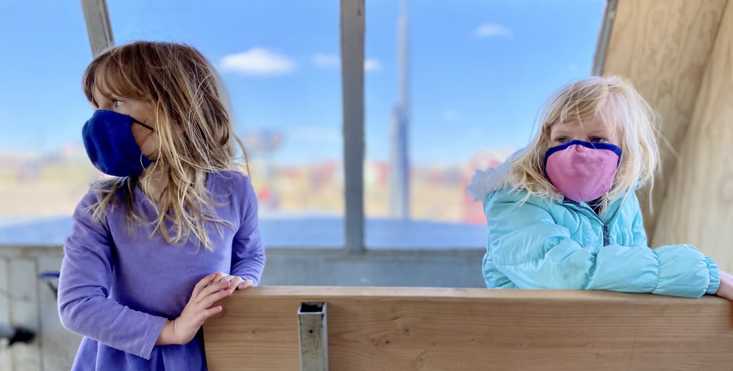 Alice, left, and Harper, right, seated on a light wooden bench looking off in the distance toward the left of the frame. Alice has long dark blonde hair and is wearing a purple dress and blue mask while Harper's light blond hair is draped over her shoulders on top of her puffy light blue jacket as she wears a pink mask with blue trim.