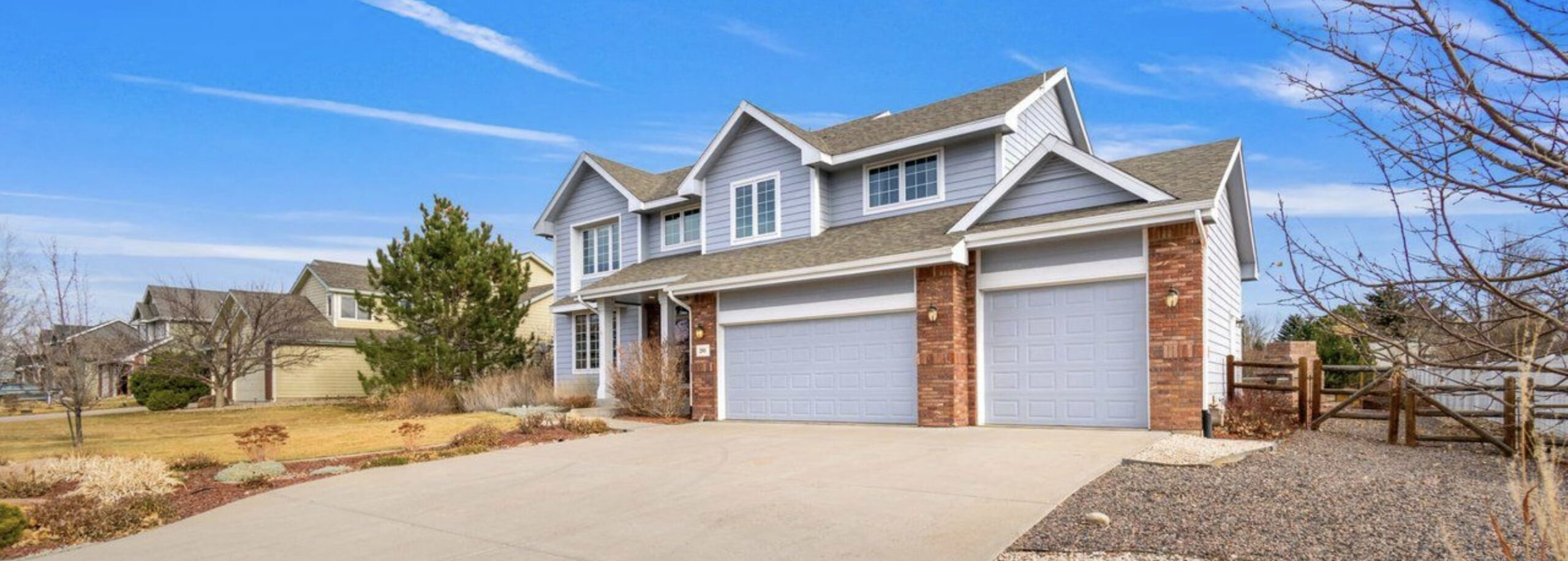 Photo of the front of a two-story house with light blue siding and brick trim against a gorgeous blue sky dotted with lazy white clouds floating overhead.